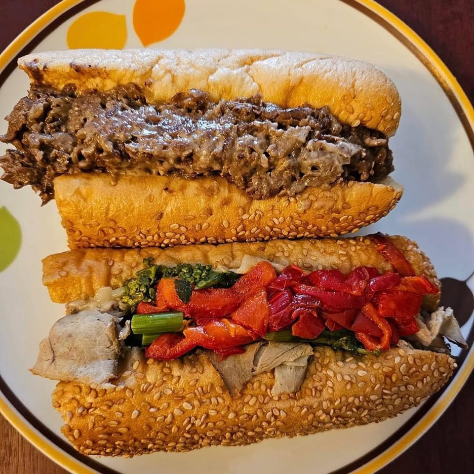 Two Brynn Bradley cheesesteaks on a plate, one with melted cheese, the other with peppers and greens.